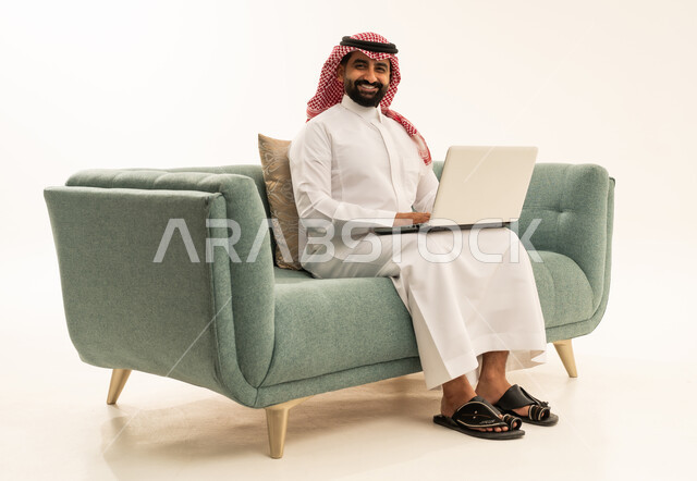 النظر للكاميرا بتعابير السرور، العمل عن بعد وانجاز المهام المطلوبة، إدارة شؤون الأعمال وتنظيمها عبر الاجهزة التقنية، دمج العمل مع التكنولوجيا، بورتريه لرجل عربي سعودي خليجي يرتدي الشماغ والثوب التقليدي يجلس على الاريكة يعمل على الحاسوب المحمول، استخدام جهاز تقني حديث ومتطور، خلفية بيضاء