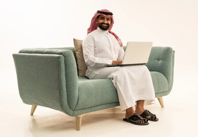 النظر للكاميرا بتعابير السرور، العمل عن بعد وانجاز المهام المطلوبة، إدارة شؤون الأعمال وتنظيمها عبر الاجهزة التقنية، دمج العمل مع التكنولوجيا، بورتريه لرجل عربي سعودي خليجي يرتدي الشماغ والثوب التقليدي يجلس على الاريكة يعمل على الحاسوب المحمول، استخدام جهاز تقني حديث ومتطور، خلفية بيضاء