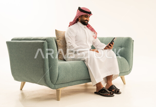 Browsing social media with gestures of joy, electronic marketing through smart devices, following the latest news on social media, a side portrait of an Arab Saudi Gulf man wearing a ghutrah and traditional thobe sitting on a sofa using a mobile phone, white background.