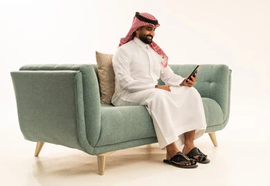 تصفح مواقع التواصل الاجتماعي بإيماءات السرور، التسويق الالكتروني عبر الأجهزة الذكية, متابعة آخر أخبار السوشال ميديا,  بورتريه من الجانب لرجل عربي سعودي خليجي يرتدي الشماغ والثوب التقليدي يجلس على الاريكة يستخدم الهاتف المحمول, خلفية بيضاء