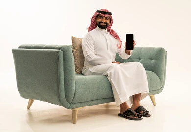 Marketing advertisements and promotional offers, display of a blank black mobile phone screen, integrating modern technologies and technology into daily life, a portrait of an Arab Saudi Gulf man wearing a ghutrah and traditional thobe sitting on a sofa holding a mobile phone in his hand and looking at the camera with a smile, full-body shot, white background.