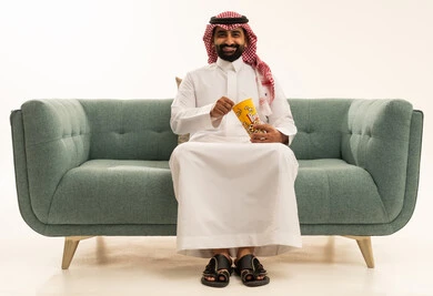 Enjoying popcorn, the concept of amusement and entertainment, expressions of joy and enjoyment, spending enjoyable and entertaining times, a portrait of an Arab Saudi Gulf man wearing a ghutrah and traditional thobe sitting on a sofa holding a box of popcorn and watching movies, watching a cinematic film, white background.