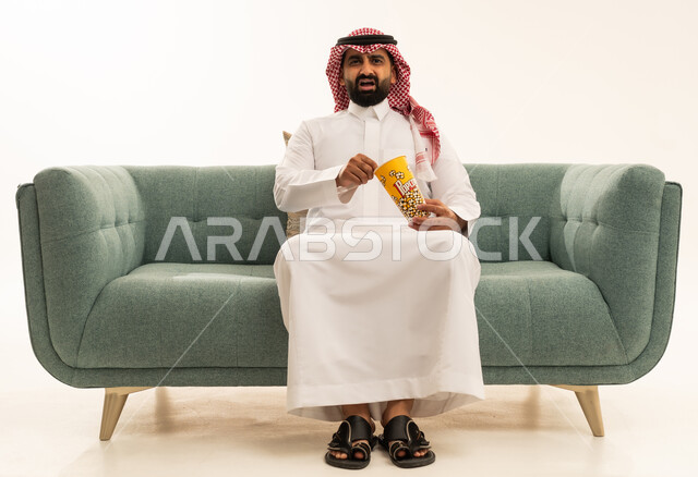 Looking with expressions of astonishment and surprise, enjoying eating popcorn, the concept of amusement and entertainment, spending enjoyable and entertaining times, a portrait of an Arab Saudi Gulf man wearing a ghutrah and traditional thobe sitting on the sofa holding a box of popcorn and watching movies, watching a cinematic film, white background.