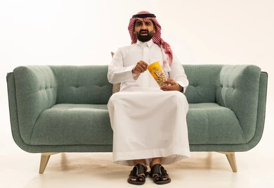 النظر بتعابير الاندهاش والاستغراب، الاستمتاع بتناول الفوشار، مفهوم التسليه والترفيه، قضاء أوقات ممتعة ومسلية، بورتريه لرجل عربي سعودي خليجي يرتدي الشماغ والثوب التقليدي يجلس على الاريكة يحمل بيده علبة الفشار ويقوم بمشاهده الافلام ، مشاهدة فيلم سينمائي ، خلفيه بيضاء