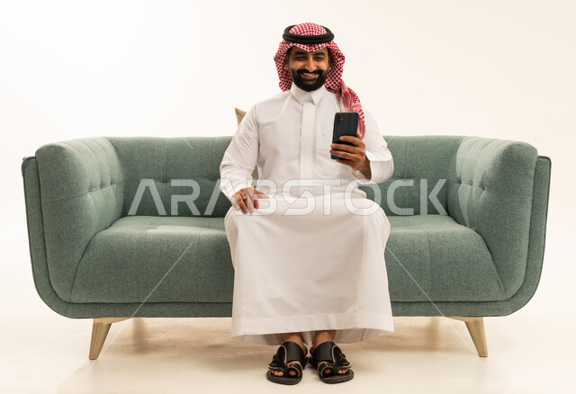 تصفح وسائل التواصل الاجتماعي عبر الجوال، استخدام الأجهزة التقنية الحديثة و المتطورة,  بورتريه لرجل عربي سعودي خليجي يرتدي الشماغ والثوب التقليدي يجلس على الاريكة يمسك الهاتف المحمول بيده, خلفية بيضاء