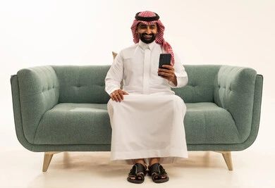 تصفح وسائل التواصل الاجتماعي عبر الجوال، استخدام الأجهزة التقنية الحديثة و المتطورة,  بورتريه لرجل عربي سعودي خليجي يرتدي الشماغ والثوب التقليدي يجلس على الاريكة يمسك الهاتف المحمول بيده, خلفية بيضاء