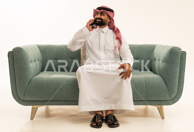 التواصل مع الاهل والاصدقاء، ايماءات السعادة والسرور لسماع الاخبار المفرحة،بورتريه لرجل عربي سعودي خليجي يرتدي الشماغ والثوب التقليدي يجلس على الاريكة يقوم باجراء مكالمة هاتفية عبر الهاتف المحمول، استخدام التقنيات والاجهزة الحديثة، صورة الجسم بالكامل، خلفية بيضاء