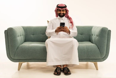 استخدام الأجهزة التقنية الحديثة و المتطورة, تصفح وسائل التواصل الاجتماعي عبر الجوال,  بورتريه لرجل عربي سعودي خليجي يرتدي الشماغ والثوب التقليدي يجلس على الاريكة يمسك الهاتف المحمول بيده, خلفية بيضاء