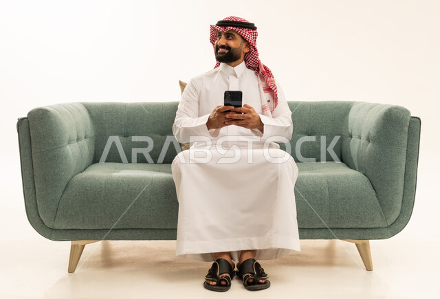 Looking at something, using modern and advanced technological devices, browsing social media via mobile, a portrait of an Arab Saudi Gulf man wearing a ghutrah and traditional thobe sitting on a sofa holding a mobile phone in his hand, white background.