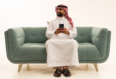 Looking at something, using modern and advanced technological devices, browsing social media via mobile, a portrait of an Arab Saudi Gulf man wearing a ghutrah and traditional thobe sitting on a sofa holding a mobile phone in his hand, white background.