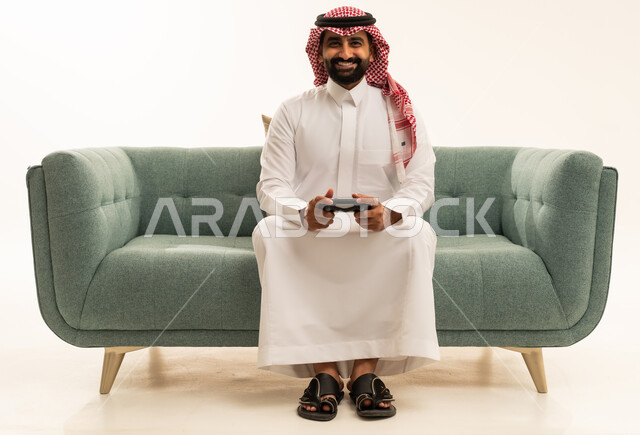 مفهوم المتعة والتسلية، إيماءات التفاعل والاندماج, بورتريه لرجل عربي سعودي خليجي يرتدي الشماغ والثوب التقليدي يجلس على الاريكة يمسك عصا التحكم ينظر إلى الكاميرا بتعابير السرور، قضاء وقت ممتع في اللعب بألعاب الفيديو، الاستمتاع بالتقنيات والتكنولوجيا الحديثة، الجسم بالطول الكامل، خلفية بيضاء
