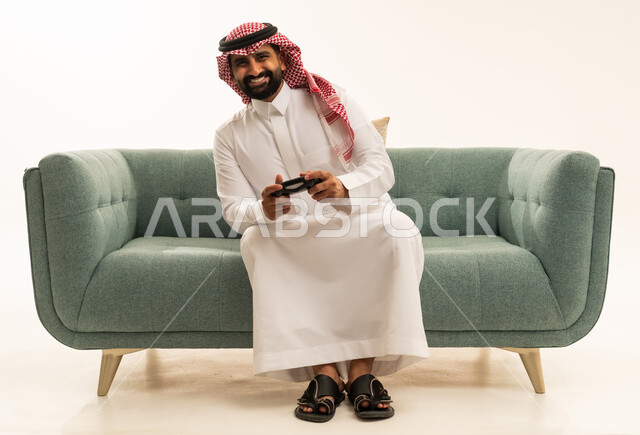إيماءات التفاعل والاندماج, بورتريه لرجل عربي سعودي خليجي يرتدي الشماغ والثوب التقليدي يجلس على الاريكة يمسك عصا التحكم ينظر إلى الكاميرا بتعابير السرور، مفهوم المتعة والتسلية، قضاء وقت ممتع في اللعب بألعاب الفيديو، الاستمتاع بالتقنيات والتكنولوجيا الحديثة، خلفية بيضاء