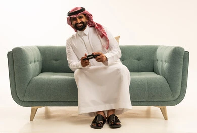 إيماءات التفاعل والاندماج, بورتريه لرجل عربي سعودي خليجي يرتدي الشماغ والثوب التقليدي يجلس على الاريكة يمسك عصا التحكم ينظر إلى الكاميرا بتعابير السرور، مفهوم المتعة والتسلية، قضاء وقت ممتع في اللعب بألعاب الفيديو، الاستمتاع بالتقنيات والتكنولوجيا الحديثة، خلفية بيضاء