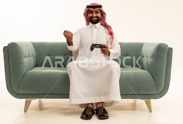 تعابير الفوز والانتصار، التفاعل والاندماج مع ألعاب البلايستيشن، تقنيات الكترونية حديثة ترفيهيه, بورتريه لرجل عربي سعودي خليجي يرتدي الشماغ والثوب التقليدي يجلس على الاريكة يحمل بيده عصا التحكم يقوم باللعب في ألعاب الفيديو، خلفية بيضاء