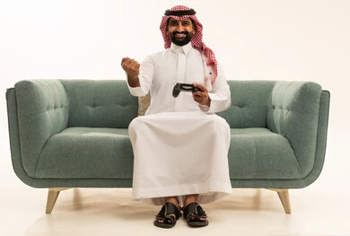 تعابير الفوز والانتصار، التفاعل والاندماج مع ألعاب البلايستيشن، تقنيات الكترونية حديثة ترفيهيه, بورتريه لرجل عربي سعودي خليجي يرتدي الشماغ والثوب التقليدي يجلس على الاريكة يحمل بيده عصا التحكم يقوم باللعب في ألعاب الفيديو، خلفية بيضاء