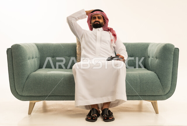 إيماءات الخسارة وخيبة الأمل، استخدام الألعاب الإلكترونية والأجهزة الحديثة، بورتريه لرجل عربي خليجي سعودي يرتدي الثوب الابيض و الشماغ يجلس على الاريكة يمسك عصا التحكم يضع يده على رأسه بتعابير الحسرة والحزن، التسلية والترفيه بالفيديو جيمز في وقت الفراغ، خلفية بيضاء
