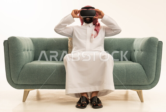 التطور التقني والتكنولوجي, المتعة و التسلية بالمشاهدة عبر نظارة الواقع الافتراضي،  بورتريه لرجل عربي خليجي سعودي يرتدي الثوب التقليدي و الشماغ يجلس على الاريكة ينظر من خلال نظارات ثلاثية الأبعاد (الفي ار), الاندماج ومحاكاة العالم الخيالي والنظر الى الافلام الواقعيه، خلفيه بيضاء