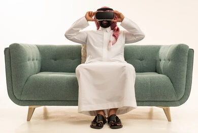 التطور التقني والتكنولوجي, المتعة و التسلية بالمشاهدة عبر نظارة الواقع الافتراضي،  بورتريه لرجل عربي خليجي سعودي يرتدي الثوب التقليدي و الشماغ يجلس على الاريكة ينظر من خلال نظارات ثلاثية الأبعاد (الفي ار), الاندماج ومحاكاة العالم الخيالي والنظر الى الافلام الواقعيه، خلفيه بيضاء