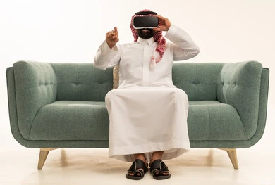 Interaction and immersion with virtual reality, pointing with the index finger at something, using modern technological devices, seeing imaginary scenes, a portrait of a Saudi Gulf Arab man wearing traditional attire and a ghutrah sitting on a sofa wearing VR glasses, spending leisure time in an enjoyable way, white background.