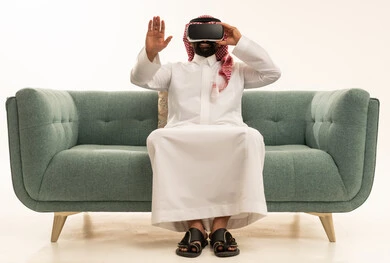 رفع كف اليد للأعلى بإيماءات الاندماج ومحاكاة العالم الخيالي، استخدام الأجهزة التقنية الحديثة، رؤية المشاهد الخيالية، بورتريه لرجل عربي خليجي سعودي يرتدي الثوب التقليدي و الشماغ يجلس على الاريكة يضع نظارات الفي آر، قضاء أوقات الفراغ بشكل ممتع، خلفية بيضاء
