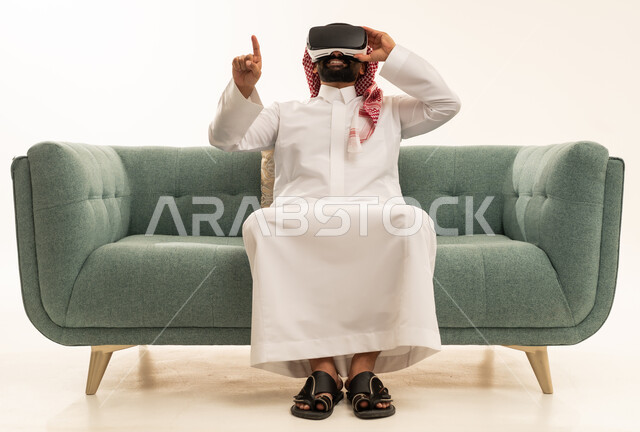 الاشارة بإصبع السبابة إلى شيء ما، استخدام الأجهزة التقنية الحديثة، التفاعل والاندماج مع الواقع الافتراضي، رؤية المشاهد الخيالية، بورتريه لرجل عربي خليجي سعودي يرتدي الثوب التقليدي و الشماغ يجلس على الاريكة يضع نظارات الفي آر، قضاء أوقات الفراغ بشكل ممتع، خلفية بيضاء