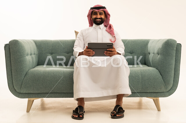 استعمال التكنولوجيا والتقنيات الحديثة، الدردشة مع الأهل والأصدقاء، بورتريه لرجل عربي سعودي خليجي يرتدي الشماغ والثوب التقليدي يجلس على الاريكة يحمل بيده جهاز التابلت، تصفح مواقع التواصل الاجتماعي، استخدام تطبيقات وبرامج الأجهزة اللوحية، خلفية بيضاء