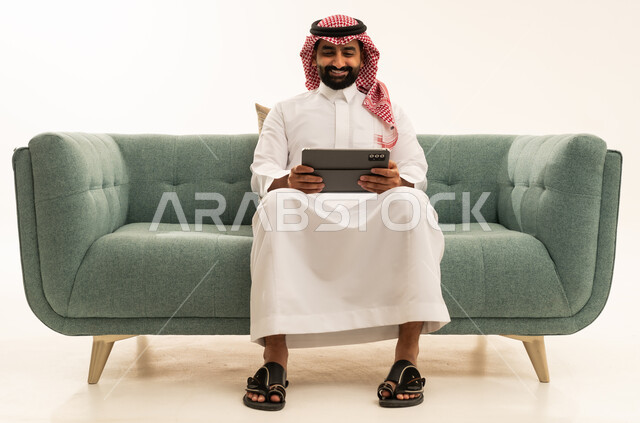 الدردشة مع الأهل والأصدقاء، استعمال التكنولوجيا والتقنيات الحديثة، بورتريه لرجل عربي سعودي خليجي يرتدي الشماغ والثوب التقليدي يجلس على الاريكة يحمل بيده جهاز التابلت، تصفح مواقع التواصل الاجتماعي، استخدام تطبيقات وبرامج الأجهزة اللوحية، خلفية بيضاء