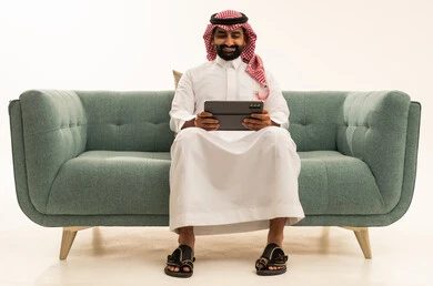 Chatting with family and friends, using technology and modern techniques, a portrait of an Arab Saudi Gulf man wearing a ghutrah and traditional thobe sitting on a sofa holding a tablet, browsing social media, using applications and programs on tablet devices, white background.