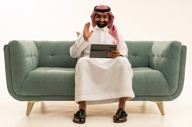 رفع كف اليد بتعابير السلام و التحية، اجراء مكالمة فيديو عبر الجهاز اللوحي، التواصل مع الاهل و الاصدقاء، بورتريه لرجل عربي سعودي خليجي يرتدي الشماغ والثوب التقليدي يجلس على الاريكة يقوم باستخدام التابلت، خلفية بيضاء