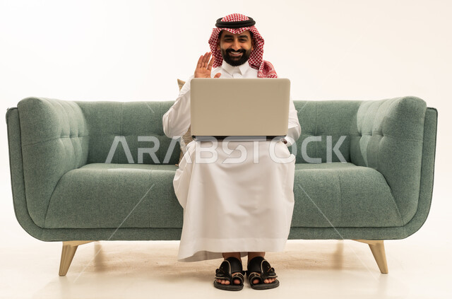 اجراء مكالمة فيديو عبر الابتوب، رفع كف اليد بتعابير السلام و التحية، التواصل مع الاهل و الاصدقاء، بورتريه لرجل عربي سعودي خليجي يرتدي الشماغ والثوب التقليدي يجلس على الاريكة يقوم باستخدام الحاسب، خلفية بيضاء