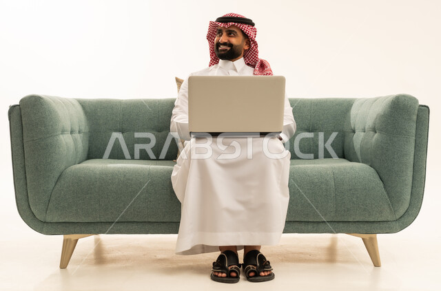 Looking at something, completing work and tasks remotely via a laptop, browsing the internet and social media, a portrait of an Arab Saudi Gulf man wearing a ghutrah and traditional thobe sitting on a sofa using a computer, white background.