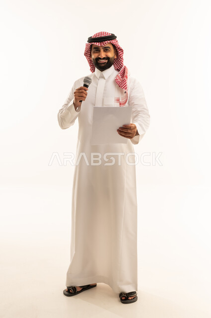 النظر للكاميرا بتعابير السرور، اعداد التقارير الخاصة بالعمل، تقديم الفقرات باستخدام الميكروفون، متابعة ومراجعة الخطط والمشاريع المكتبية، بورتريه لرجل عربي خليجي سعودي يرتدي الثوب التقليدي والشماغ  ينظر الى الاوراق البيضاء يمسك بيده المايك، الجسم بالطول الكامل، خلفية بيضاء