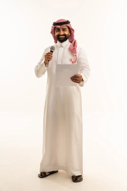 Looking at the camera with expressions of joy, preparing work-related reports, presenting segments using a microphone, following up and reviewing office plans and projects, a portrait of a Saudi Arabian Gulf man wearing traditional attire and a ghutrah, looking at white papers while holding a microphone in his hand, full-body shot, white background.