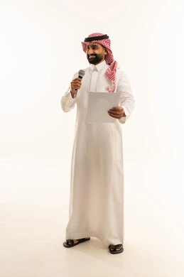 تقديم الفقرات باستخدام الميكروفون ، اعداد التقارير الخاصة بالعمل، متابعة ومراجعة الخطط والمشاريع المكتبية، بورتريه لرجل عربي خليجي سعودي يرتدي الثوب التقليدي والشماغ  ينظر الى الاوراق البيضاء يمسك بيده المايك, التدقيق في قراءة الملاحظات، الجسم بالطول الكامل، خلفية بيضاء