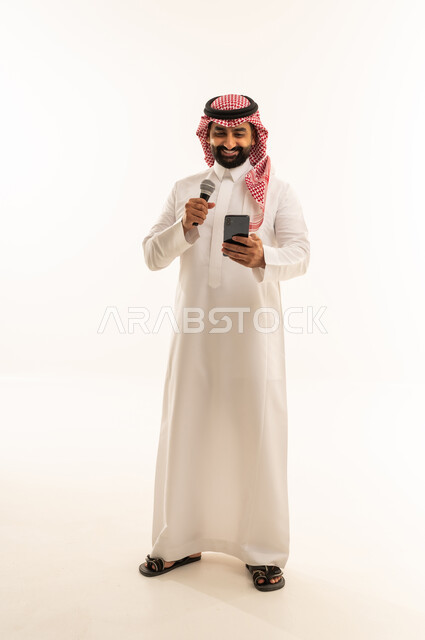 الفن المسرحي الغنائي، ممارسة هواية الغناء للتسلية والترفيه، مواهب فنية في عالم الموسيقى، تقديم العرض المسرحي، بورتريه لرجل عربي خليجي سعودي يرتدي الثوب و الشماغ  يحمل بيده الميكروفون و الهاتف المحمول ، الجسم بالطول الكامل، خلفية بيضاء