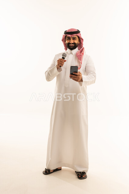 Practicing the hobby of singing for fun and entertainment, artistic talents in the world of music, musical theater art, presenting the theatrical performance, a portrait of an Arab Gulf Saudi man wearing a thobe and a ghutrah holding a microphone and a mobile phone in his hand, full-body, white background.