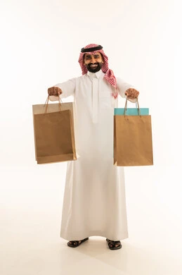 Shopping for supplies and shopping at commercial malls, taking advantage of seasonal offers and discounts, a portrait of a Saudi Arabian Gulf man wearing a thobe and a ghutrah holding colorful paper bags in his hands and looking at the camera, full body, white background.