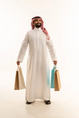 الاستفادة من العروض الموسمية و التخفيضات، شراء المستلزمات و التبضع من المولات التجارية, بورتريه لرجل عربي خليجي سعودي يرتدي الثوب و الشماغ يمسك بيدية أكياس ورقية ملونة و ينظر الى الكاميرا، الجسم بالطول الكامل، خلفية بيضاء