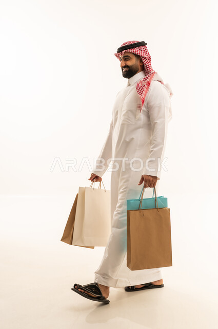 التبضع وأخذ الحاجات اللازمة والضرورية, المشتريات التجارية والهدايا, مواسم التخفيضات في السعودية, الاستفادة من الحسومات والتصفيات, بورتريه من الجانب لرجل عربي خليجي سعودي يرتدي الثوب و الشماغ يمسك بيده أكياس ورقية, متعة التسوق في المملكة, جسم بالطول الكامل, خلفية بيضاء