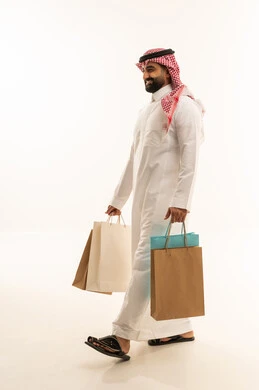 التبضع وأخذ الحاجات اللازمة والضرورية, المشتريات التجارية والهدايا, مواسم التخفيضات في السعودية, الاستفادة من الحسومات والتصفيات, بورتريه من الجانب لرجل عربي خليجي سعودي يرتدي الثوب و الشماغ يمسك بيده أكياس ورقية, متعة التسوق في المملكة, جسم بالطول الكامل, خلفية بيضاء