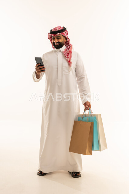 تصفح الانترنت عبر الجوال، مفهوم التسوق عن بعد, عمليات الشراء الالكتروني عبر الانترنت, بورتريه لرجل عربي خليجي سعودي يرتدي الثوب و الشماغ يمسك بيدية أكياس ورقية ملونة، التبضع والاستفادة من العروض والتخفيضات, الجسم بالطول الكامل, خلفية بيضاء