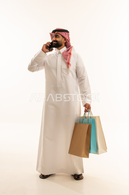 اجراء مكالمة هاتفية عبر الجوال، مفهوم التسوق عن بعد, عمليات الشراء الالكتروني عبر الانترنت, بورتريه لرجل عربي خليجي سعودي يرتدي الثوب و الشماغ يمسك بيدية أكياس ورقية ملونة، التبضع والاستفادة من العروض والتخفيضات, الجسم بالطول الكامل, خلفية بيضاء