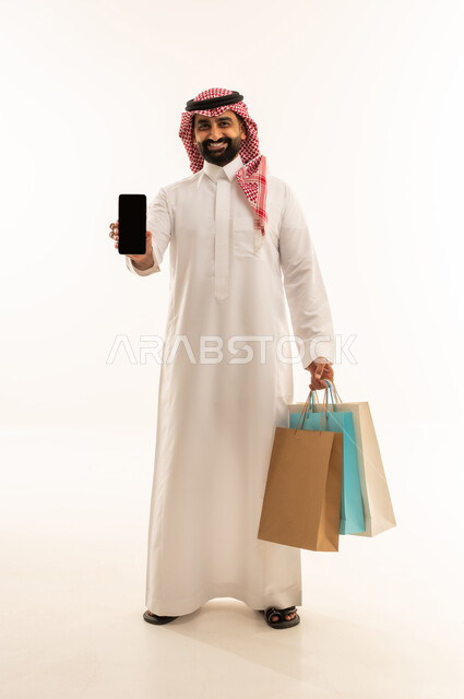 عرض شاشة سوداء فارغه عبر الجوال، مفهوم التسوق عن بعد, عمليات الشراء الالكتروني عبر الانترنت, بورتريه لرجل عربي خليجي سعودي يرتدي الثوب و الشماغ يمسك بيدية أكياس ورقية ملونة، التبضع والاستفادة من العروض والتخفيضات, الجسم بالطول الكامل, خلفية بيضاء