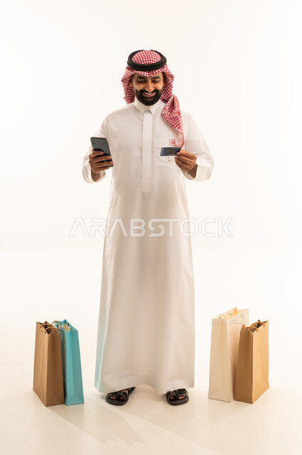 الشراء والدفع عن طريق بطاقة الإئتمان البنكية، إتمام عملية الطلبات باستخدام الهاتف المحمول، مفهوم التسوق عن بعد, بورتريه لرجل عربي خليجي سعودي يرتدي الثوب التقليدي والشماغ يقف بجانب حقائب ورقية يمسك بيده الجوال و كارت بنكي , التبضع والاستفادة من العروض والتخفيضات, الجسم بالطول الكامل، خلفية بيضاء