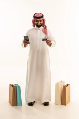 Purchasing and payment via bank credit card, completing order processes using mobile phones, the concept of remote shopping, a portrait of a Saudi Arabian Gulf man wearing traditional attire and a ghutrah standing next to paper bags holding a mobile phone and a bank card, shopping and benefiting from offers and discounts, full-body, white background.