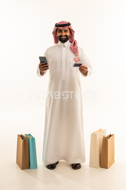مفهوم التسوق عن بعد، الشراء والدفع عن طريق بطاقة الإئتمان البنكية، إتمام عملية الطلبات باستخدام الهاتف المحمول, بورتريه لرجل عربي خليجي سعودي يرتدي الثوب التقليدي والشماغ يقف بجانب حقائب ورقية يمسك بيده الجوال و كارت بنكي , التبضع والاستفادة من العروض والتخفيضات, الجسم بالطول الكامل، خلفية بيضاء