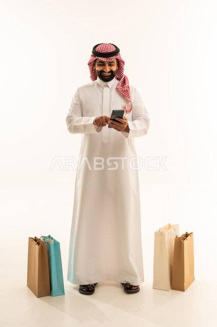 Completing the ordering process using a mobile phone, the concept of remote shopping, online electronic purchases, a portrait of an Arab Gulf Saudi man wearing a thobe and a ghutrah standing next to colorful paper bags holding a mobile phone in his hands, shopping and benefiting from offers and discounts, full-body, white background.
