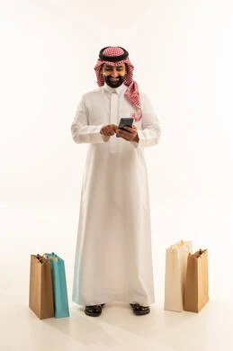 إتمام عملية الطلبات باستخدام الهاتف المحمول، مفهوم التسوق عن بعد, عمليات الشراء الالكتروني عبر الانترنت, بورتريه لرجل عربي خليجي سعودي يرتدي الثوب و الشماغ  يقف بجانب أكياس ورقية ملونة يمسك بيدية الجوال, التبضع والاستفادة من العروض والتخفيضات, الجسم بالطول الكامل, خلفية بيضاء