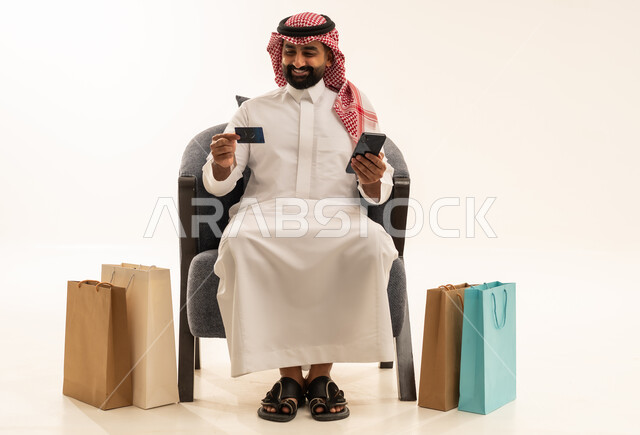 إتمام عملية الطلبات باستخدام الهاتف المحمول، الشراء والدفع عن طريق بطاقة الإئتمان البنكية، مفهوم التسوق عن بعد, بورتريه لرجل عربي خليجي سعودي يرتدي الثوب التقليدي والشماغيجلس على كرسي بجانبة أكياس ورقية ملونة, التبضع والاستفادة من العروض والتخفيضات, خلفية بيضاء
