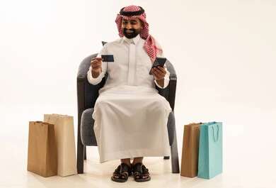 إتمام عملية الطلبات باستخدام الهاتف المحمول، الشراء والدفع عن طريق بطاقة الإئتمان البنكية، مفهوم التسوق عن بعد, بورتريه لرجل عربي خليجي سعودي يرتدي الثوب التقليدي والشماغيجلس على كرسي بجانبة أكياس ورقية ملونة, التبضع والاستفادة من العروض والتخفيضات, خلفية بيضاء